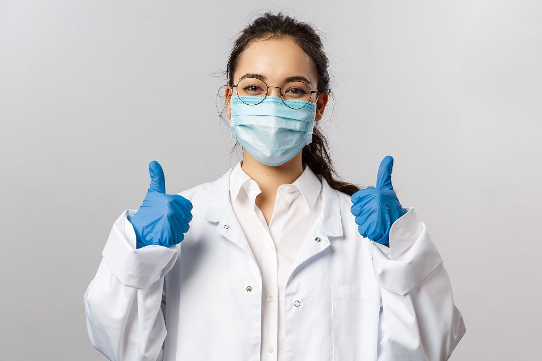 Person wearing a white coat, face mask, and gloves giving a thumbs-up gesture on a gray background